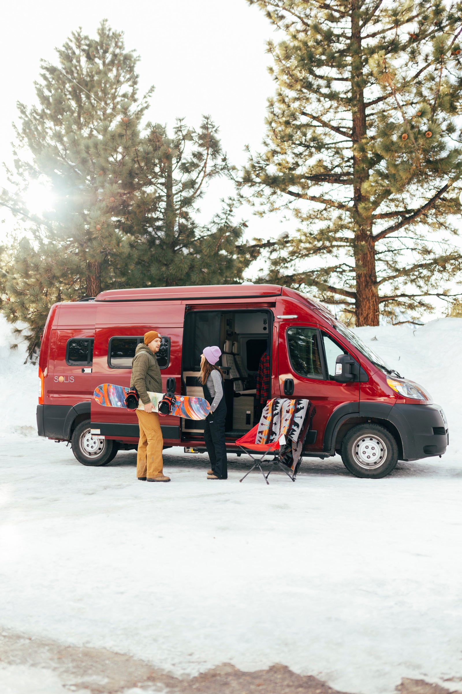 Snowy RV rental trip: Man with snowboard, woman by red camper van, chair, blanket in winter scene.