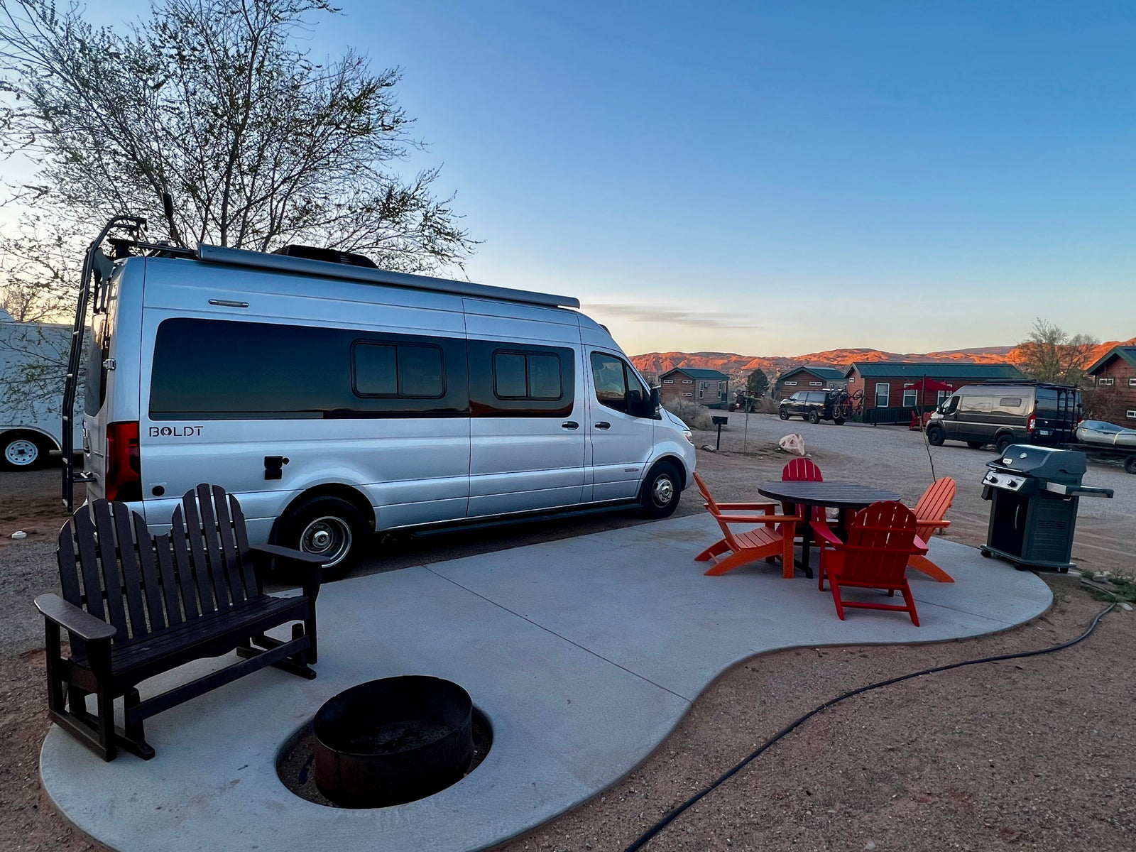 Silver Boldt campervan RV rental setup at a scenic campsite, complete with a patio, fire pit, and BBQ grill.