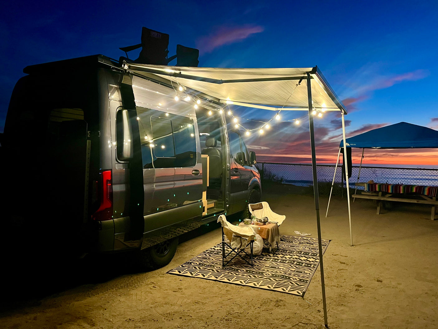 An RV rental with an illuminated awning creates a cozy evening scene by the ocean. String lights glow over a rug and chairs.