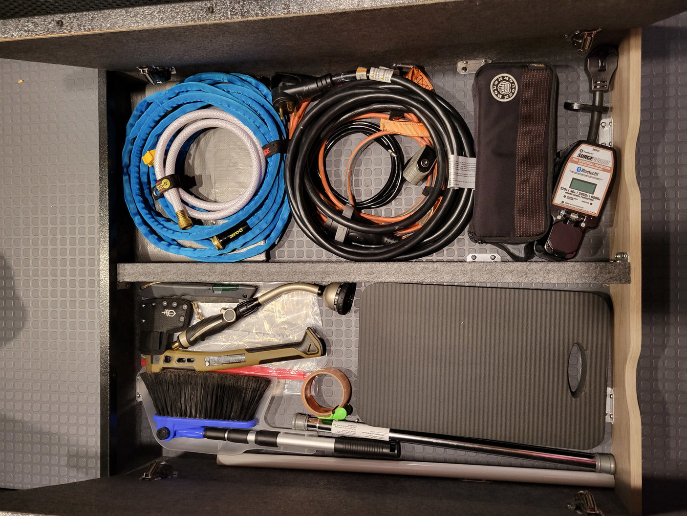 Organized RV rental compartment: water hose, power cords, Surge Guard electrical protection, and various tools neatly stored for travel.