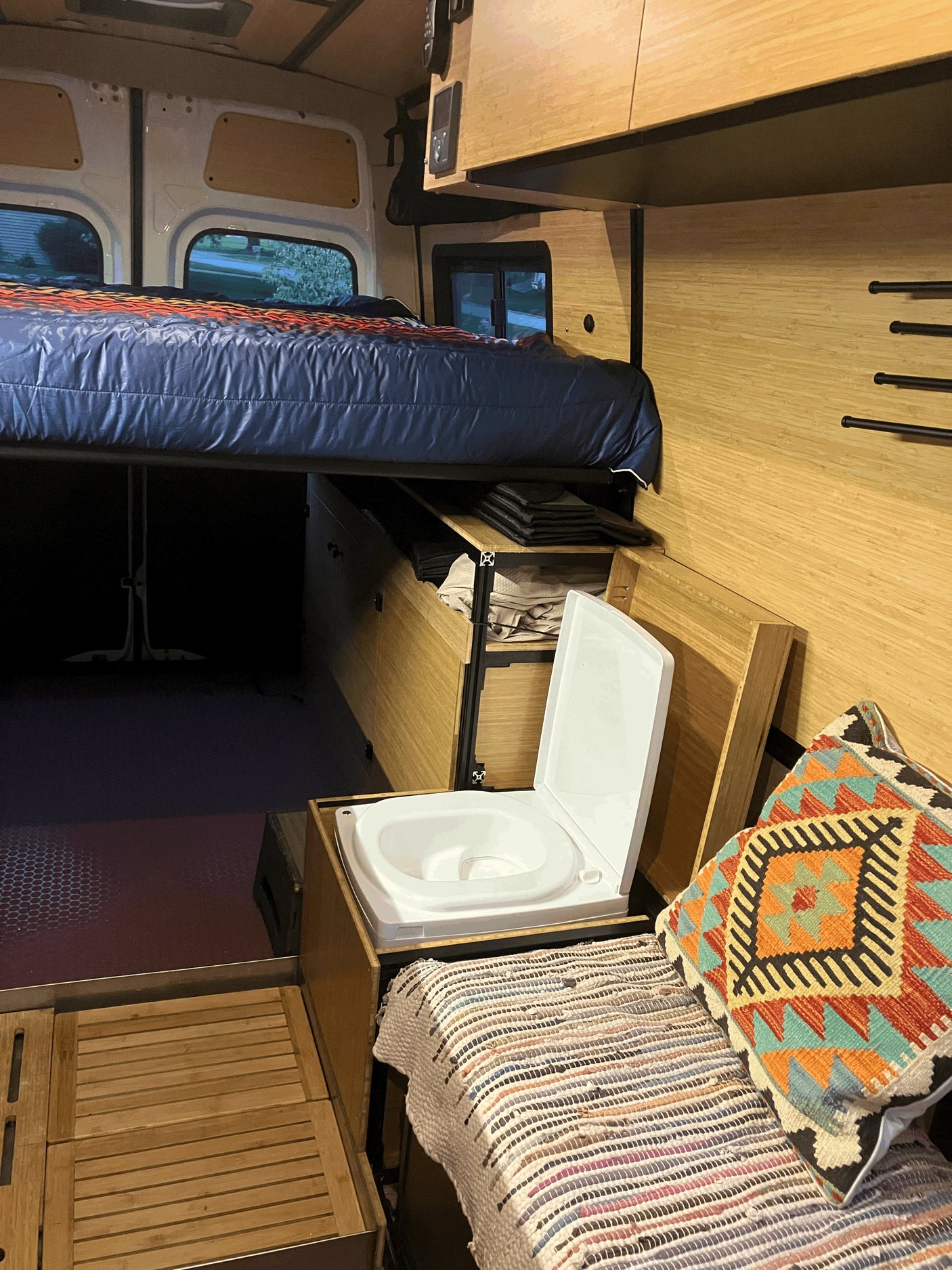 Inside an RV rental, a compact interior offers an elevated bed, bamboo-paneled storage, and a white portable toilet.