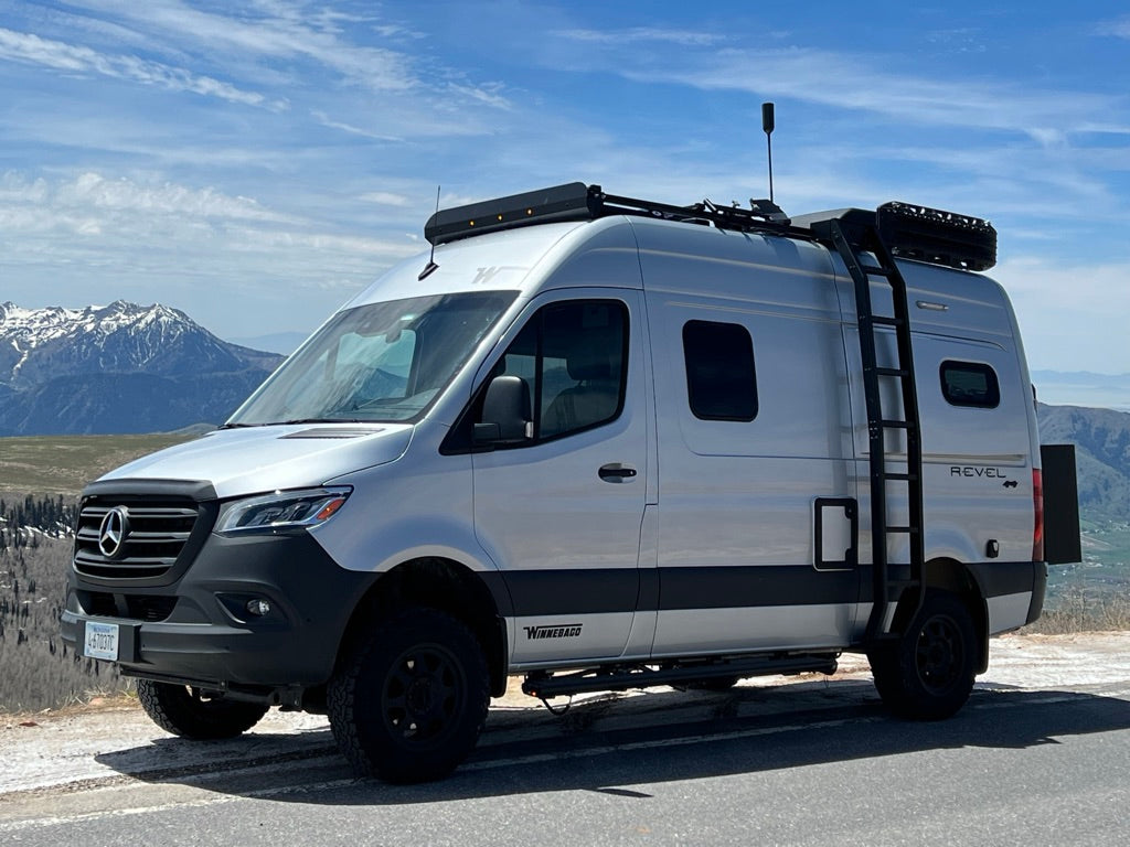 This silver Winnebago Revel 4x4 camper van, ideal for rv rental adventures, is parked on a mountain road with snowy peaks.