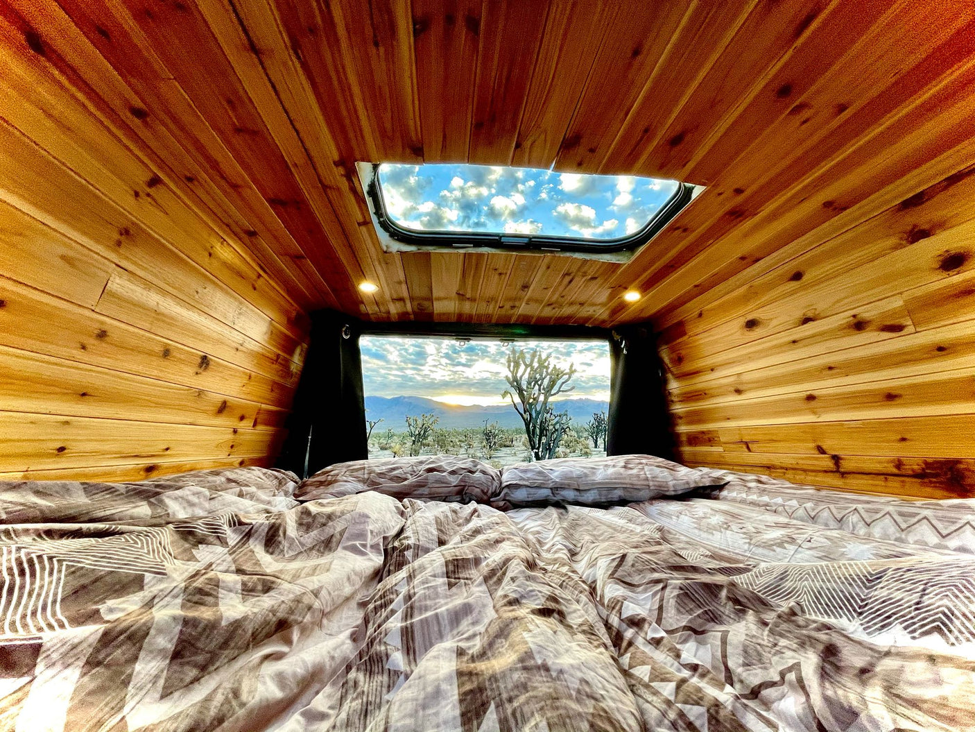 RV rental bedroom with wood walls, ceiling skylight & desert view of Joshua trees at sunrise.