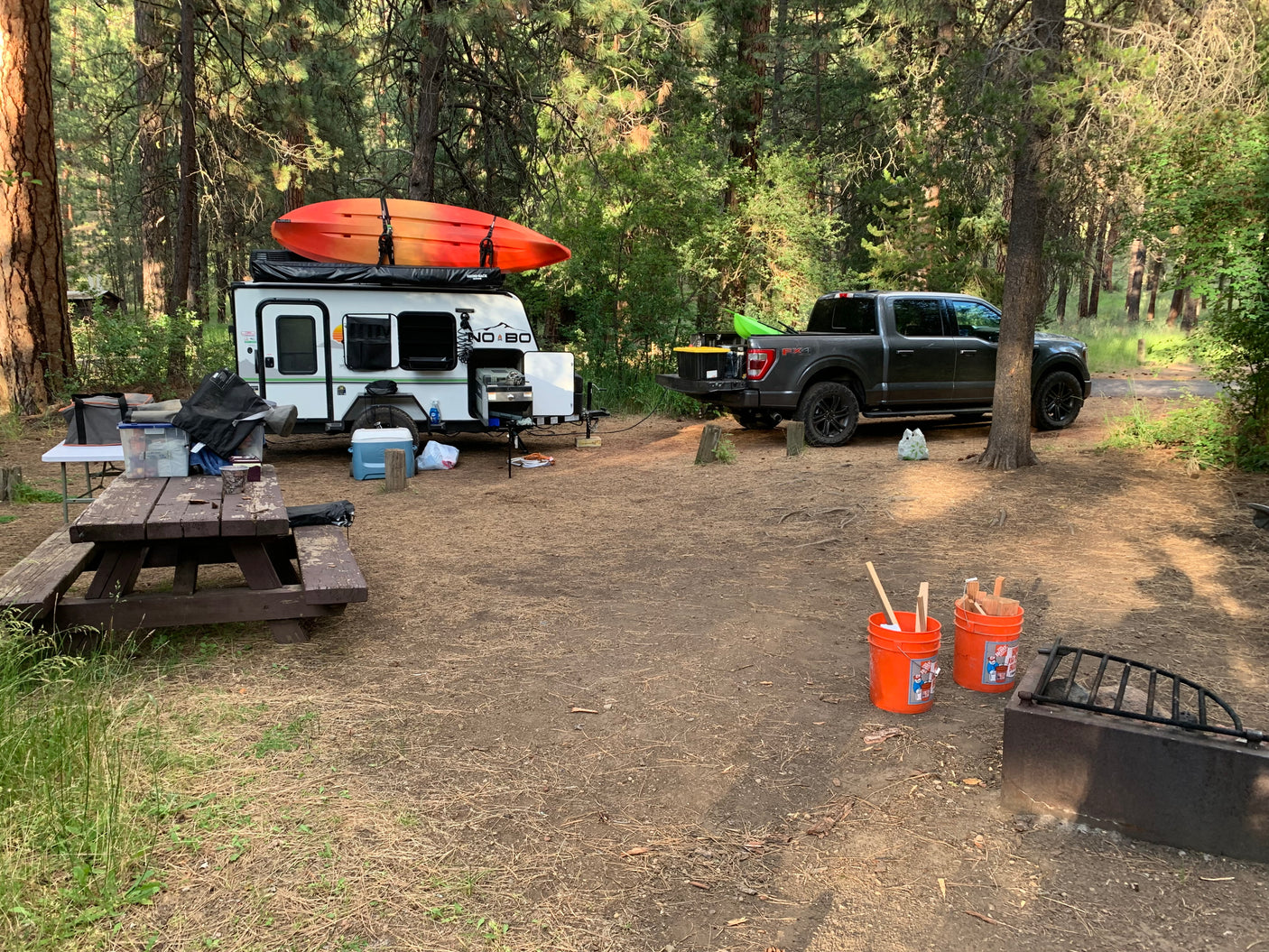 RV rental camper trailer, pickup truck, and kayaks parked at a spacious forest campsite.