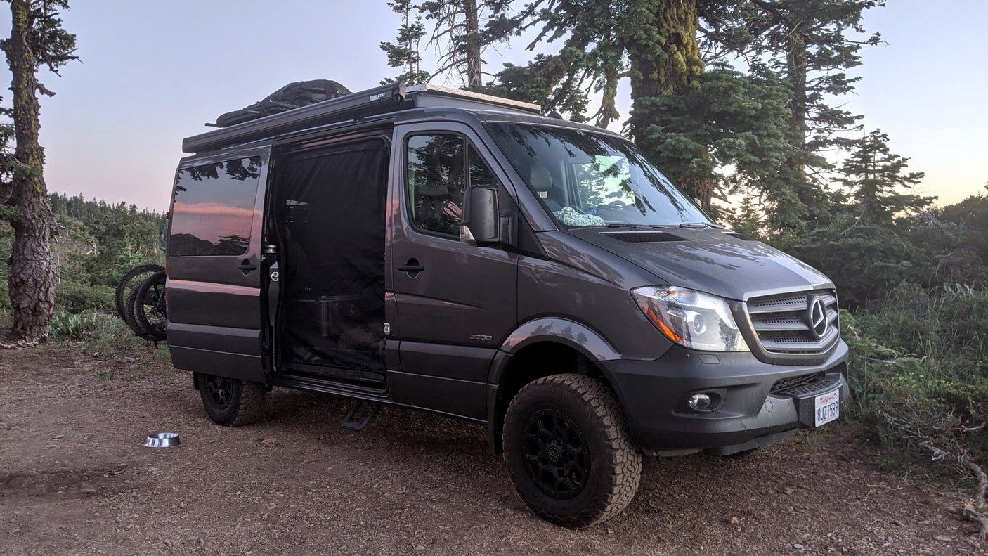 A custom dark gray Mercedes Sprinter van, perfect for an rv rental, parked on a dirt trail in a forest with an open side door.