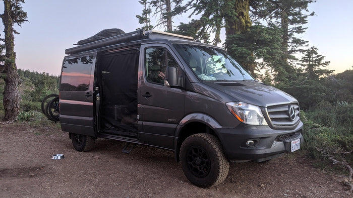 A custom dark gray Mercedes Sprinter van, perfect for an rv rental, parked on a dirt trail in a forest with an open side door.