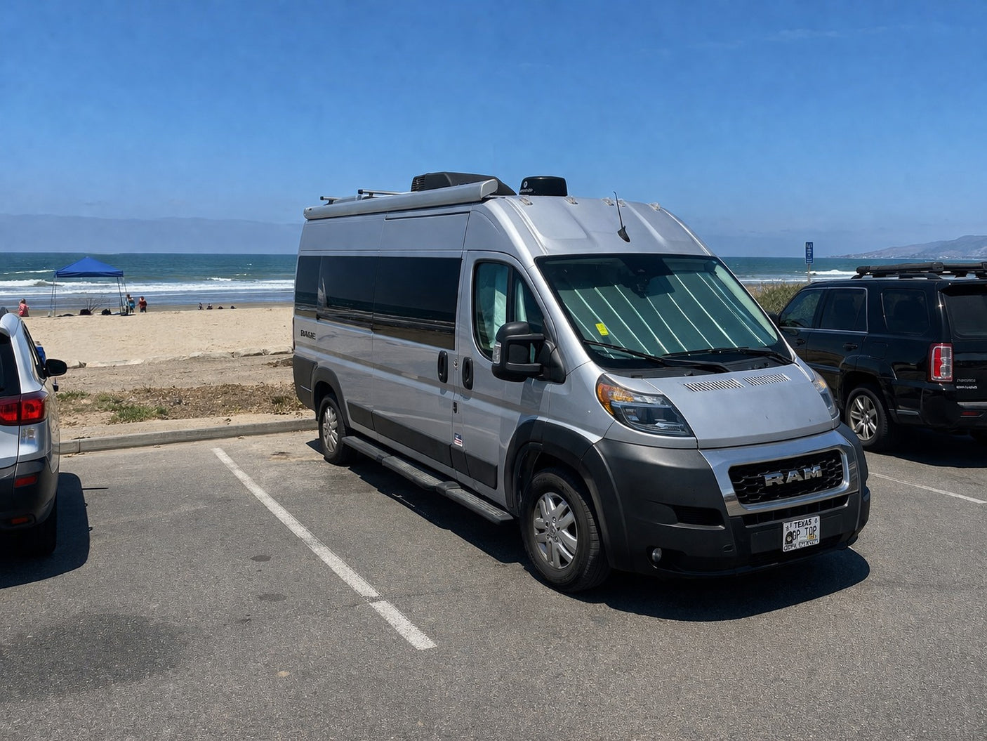 Ram ProMaster RV rental parked at a sunny beach. Ocean, mountains, & people under a blue canopy in the background.