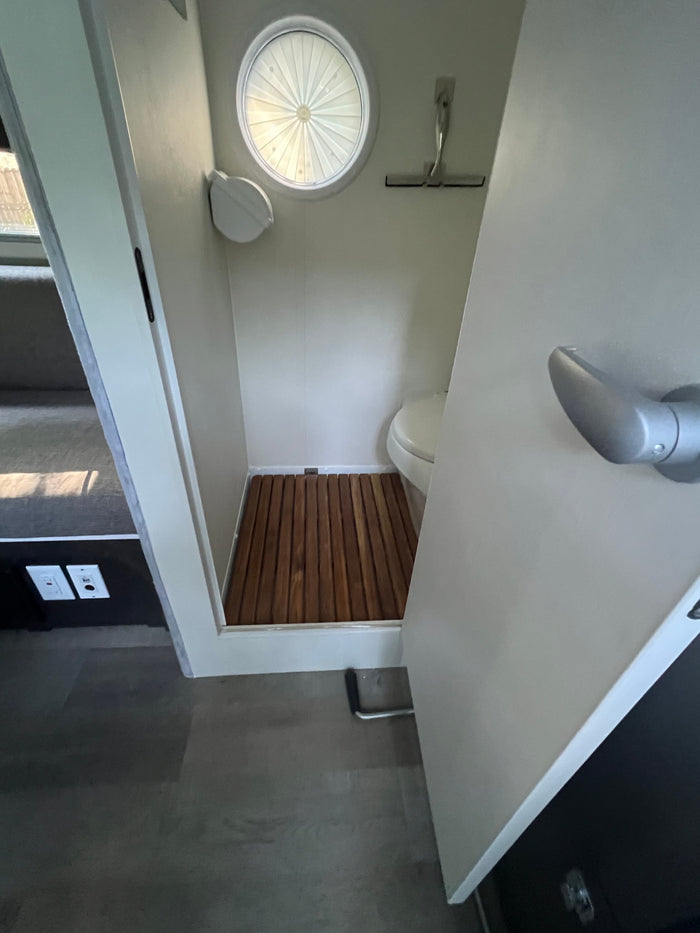RV rental wet bath, showing compact toilet, wooden shower floor, and circular window with pleated shade.