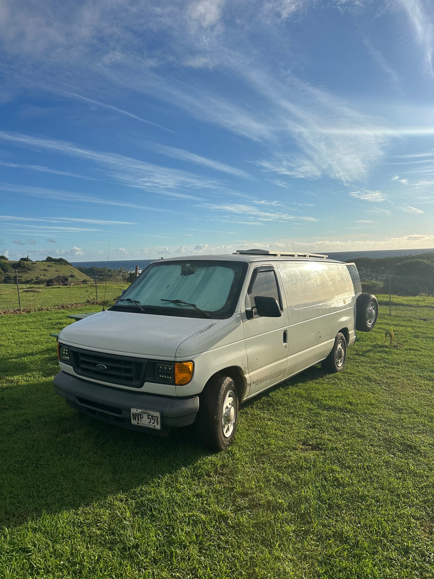 Scenic Hawaii rv rental: A white camper van parked on a grassy field overlooking the ocean under a blue sky.
