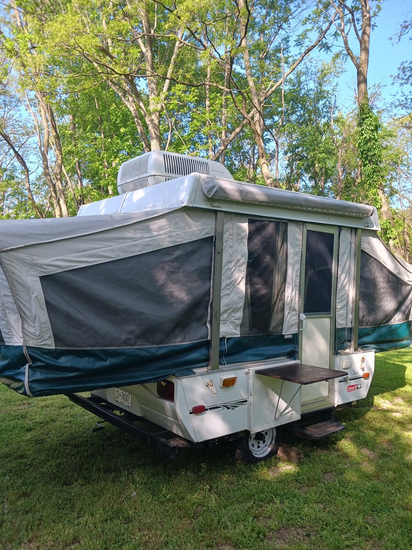 This white and green pop-up camper RV rental, with an AC unit, is set up on a grassy campsite under lush trees.