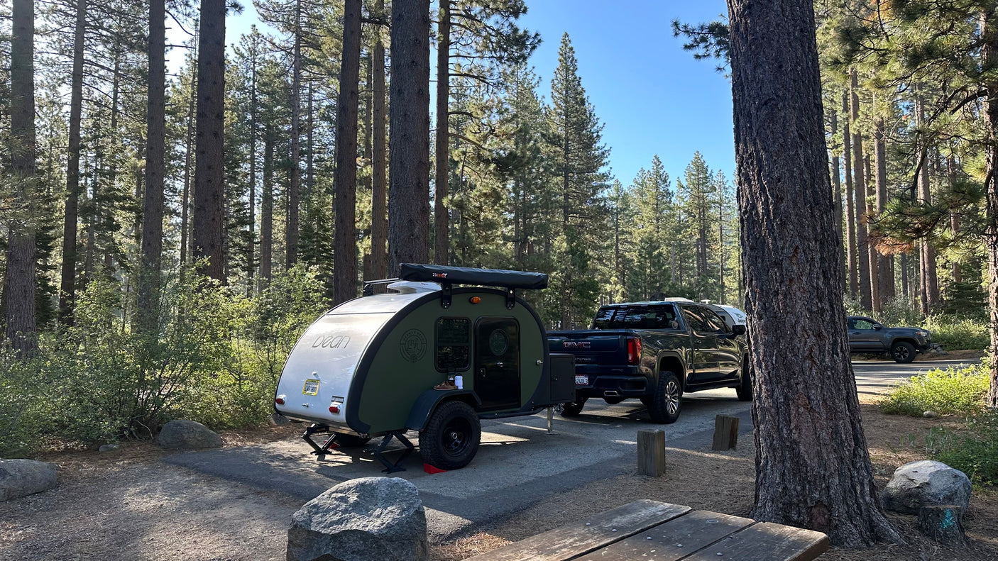 A distinctive green Bean teardrop RV rental with a GMC pickup truck, parked in a sunny pine forest campground.