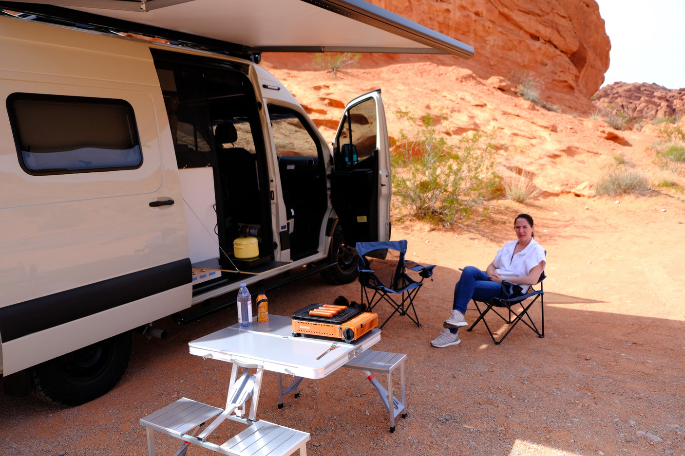 Enjoying desert camping with an **RV rental**. A woman relaxes by a camper van, grilling hot dogs on a portable stove.