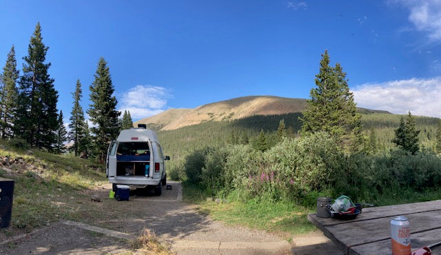 RV rental adventure! Camper van parked amidst tall trees, with open back doors, facing majestic mountains & forest. Ideal for camping.