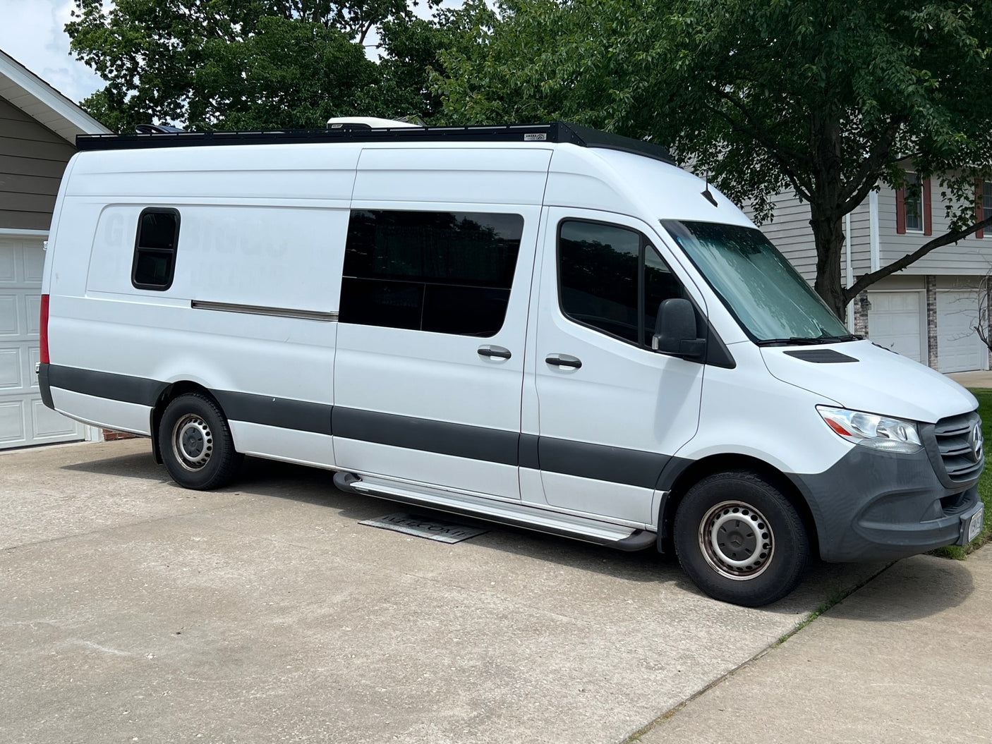 Side view of a white Mercedes Sprinter RV rental van, parked on concrete. Features tinted windows, roof AC, and side steps.