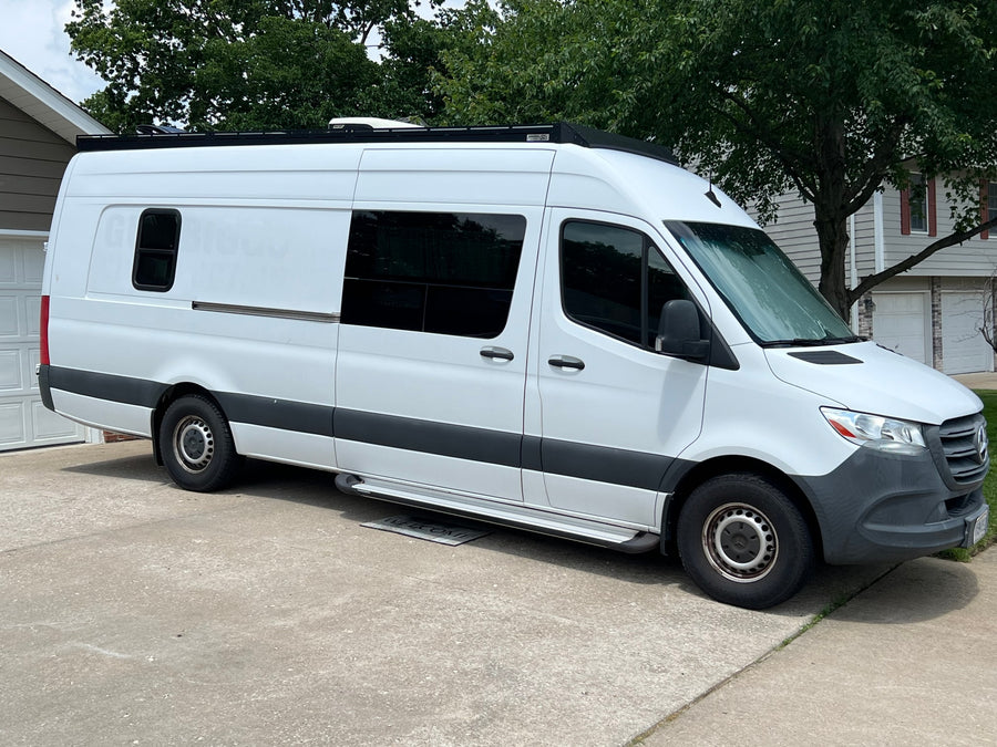 Side view of a white Mercedes Sprinter RV rental van, parked on concrete. Features tinted windows, roof AC, and side steps.
