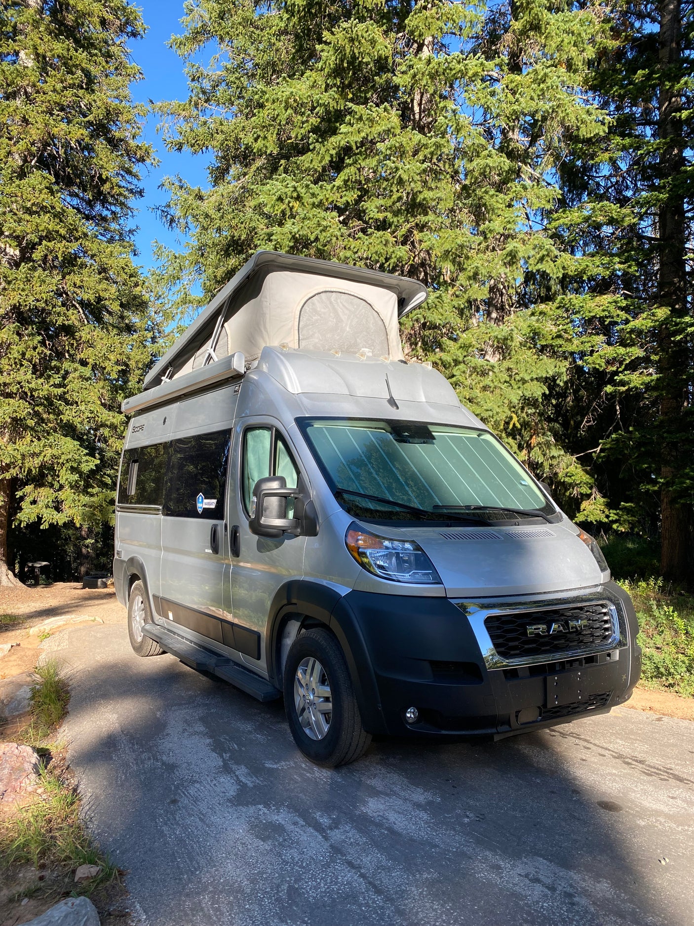 This silver pop-top RV rental, a camper van, is parked in a sunny wooded campsite, ready for adventure.