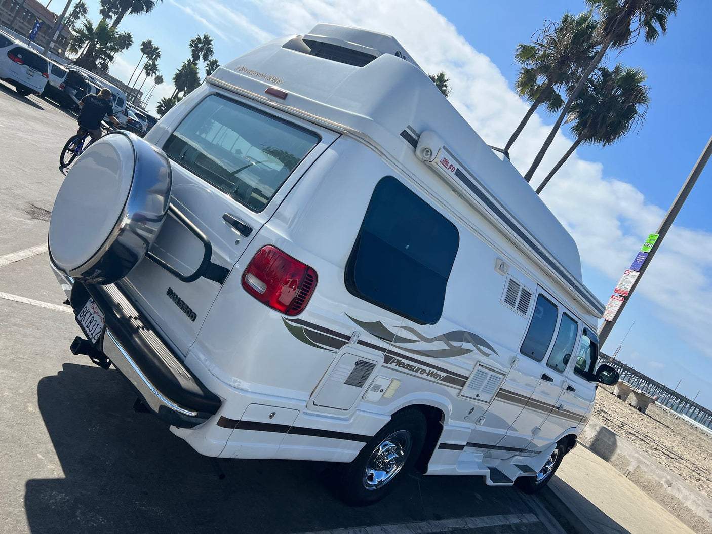White Pleasure-Way RV rental, featuring a spare tire & graphics, parked on a sunny day near a beach and pier.