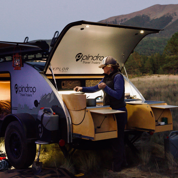 A person cooks in their Pindrop teardrop RV rental's outdoor kitchen at dusk, against a mountain backdrop.