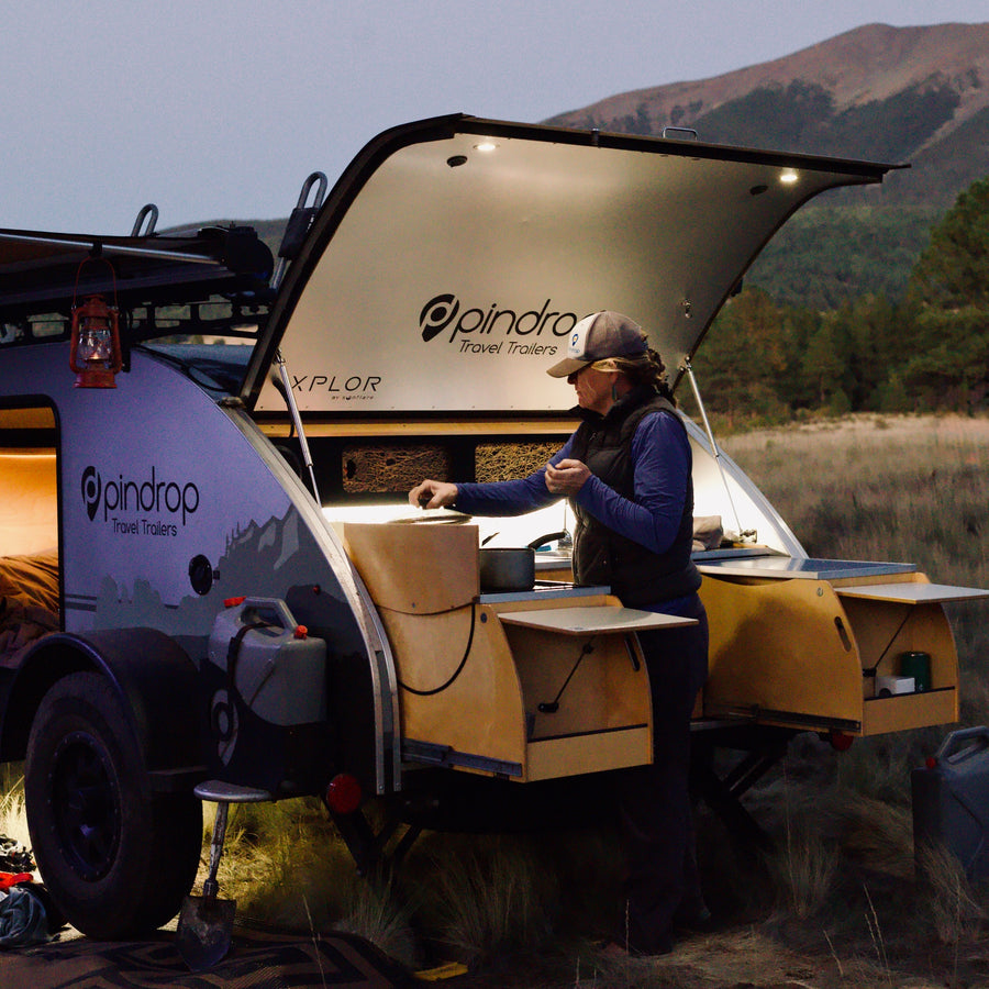 A person cooks in their Pindrop teardrop RV rental's outdoor kitchen at dusk, against a mountain backdrop.