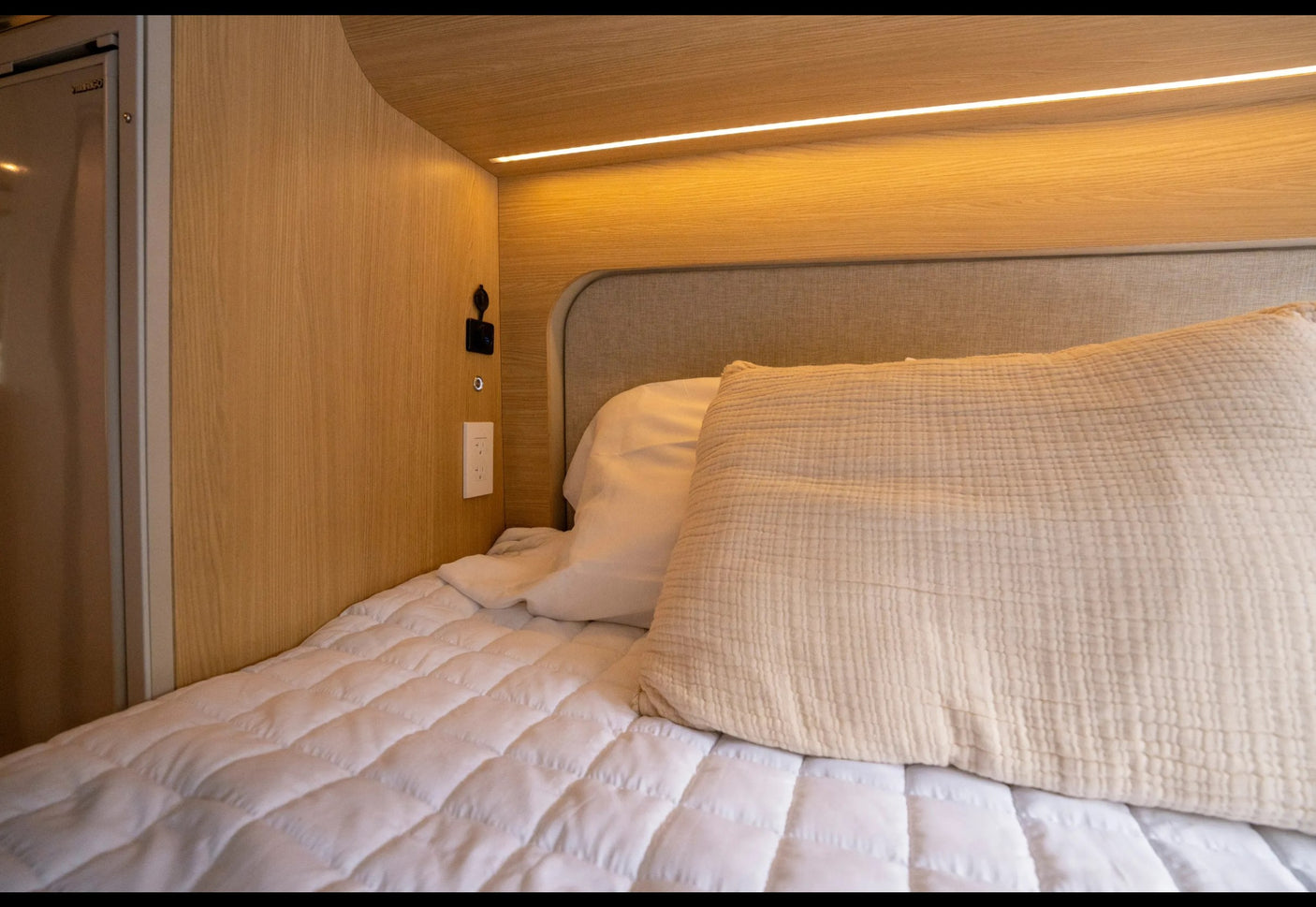 A cozy **RV rental** bed features a white quilt, textured pillow, wood paneling, and warm LED lighting with outlets.