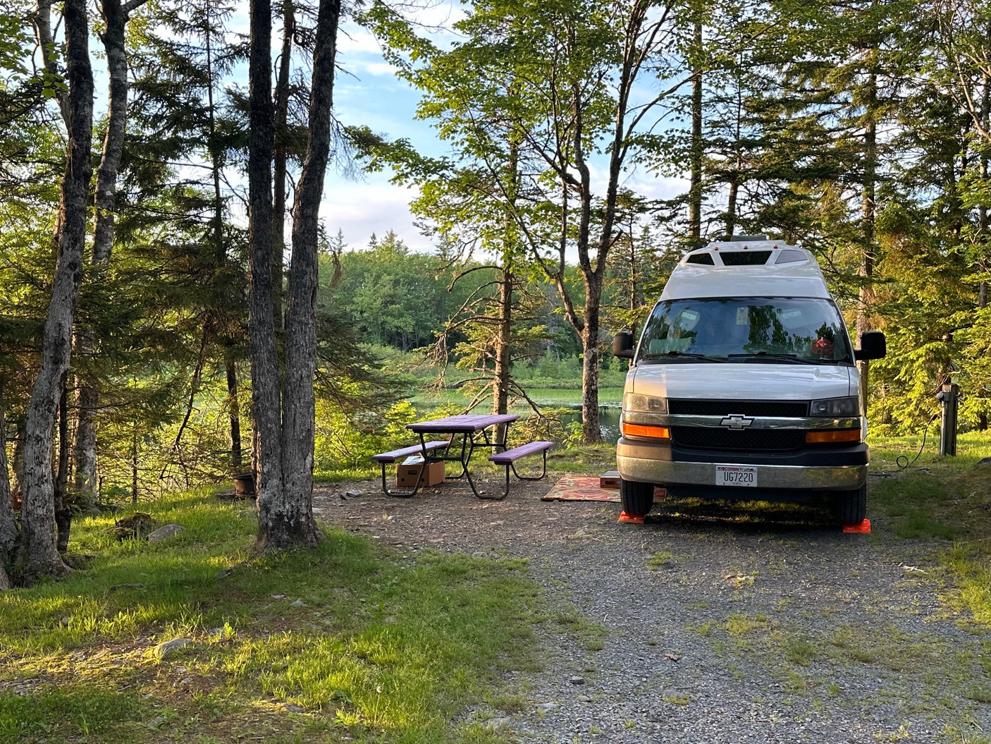 RV rental parked at a serene, wooded campsite by a lake, featuring a purple picnic table and leveling blocks under the van.