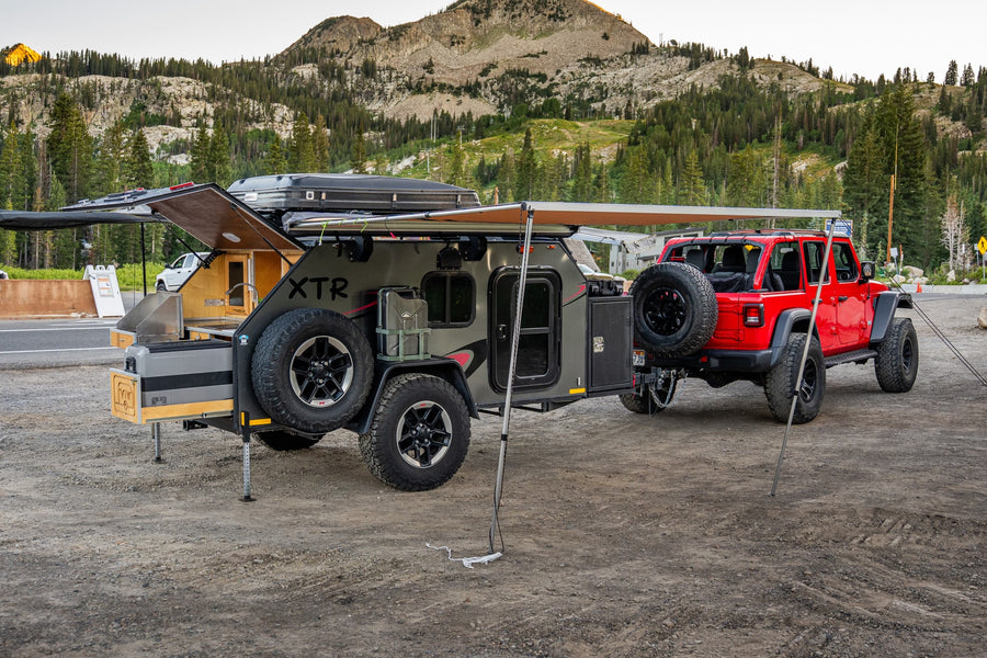 This rv rental setup features a red Jeep and a grey overlanding trailer with an extended awning and kitchen, ready for mountain camping.