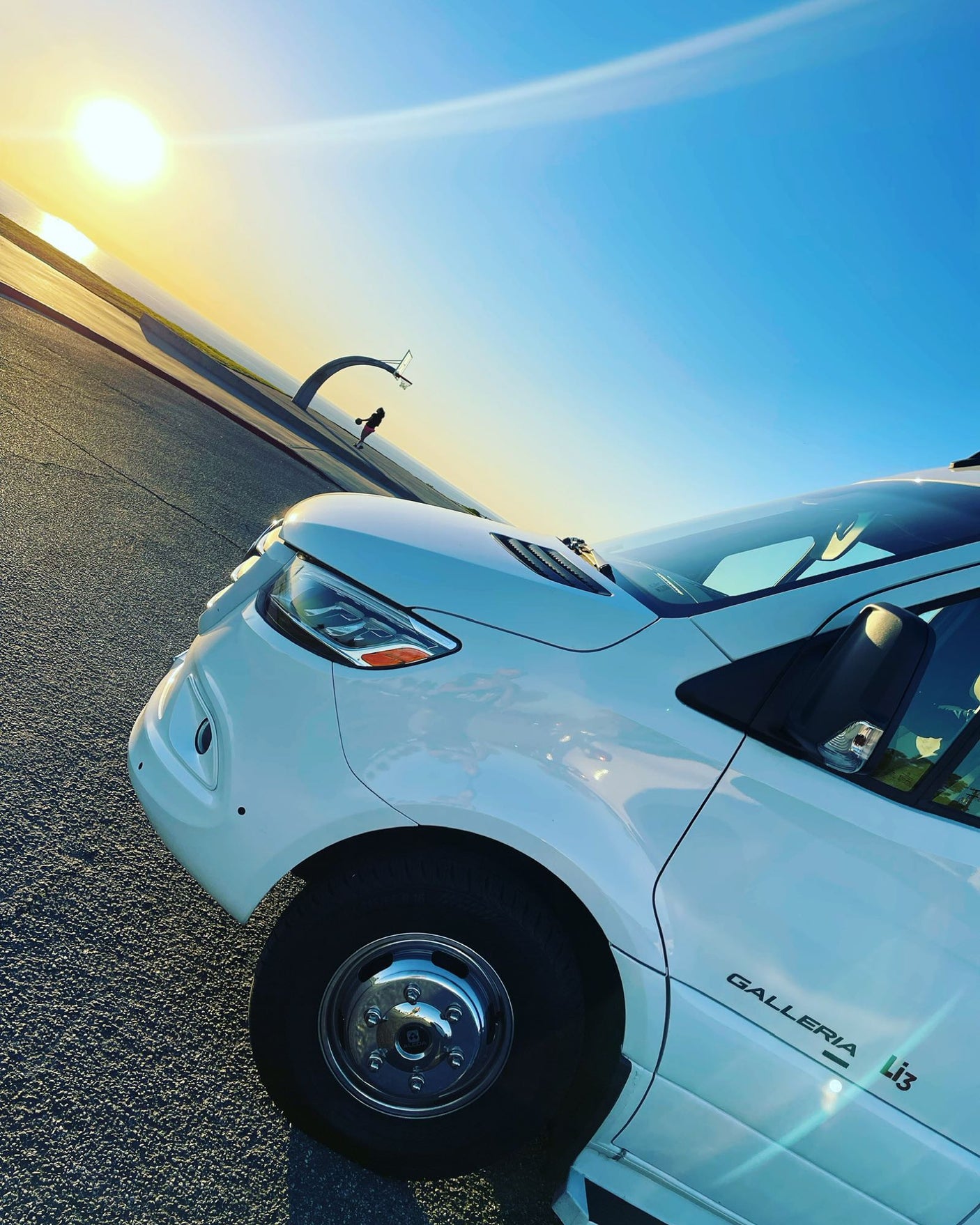White RV rental at a scenic ocean view basketball court during sunset, with a person playing in the background.