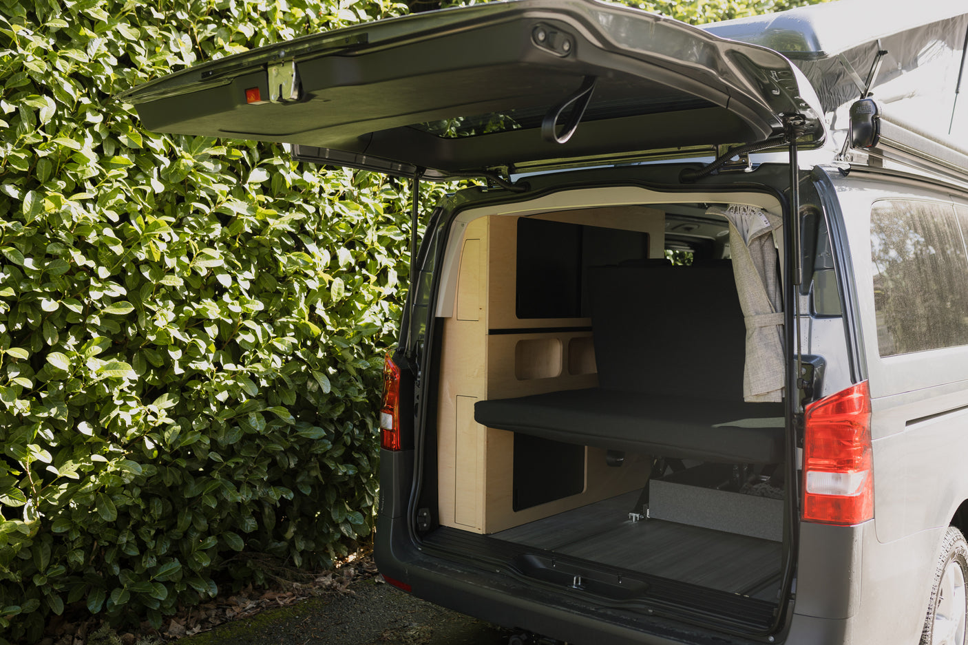 Rear view of an RV rental campervan with its tailgate open, revealing a custom wooden interior and a dark grey mattress/bench.