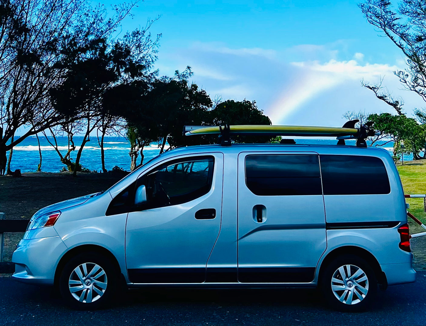 Scenic rv rental parked by the ocean with a surfboard on top & rainbow. Perfect for coastal adventures.