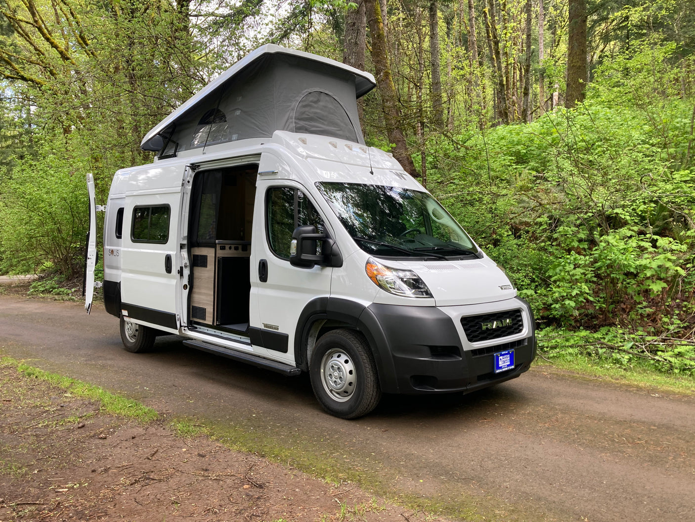 Explore the outdoors in this white pop-top rv rental van with its side door open, parked on a dirt road in a green forest.
