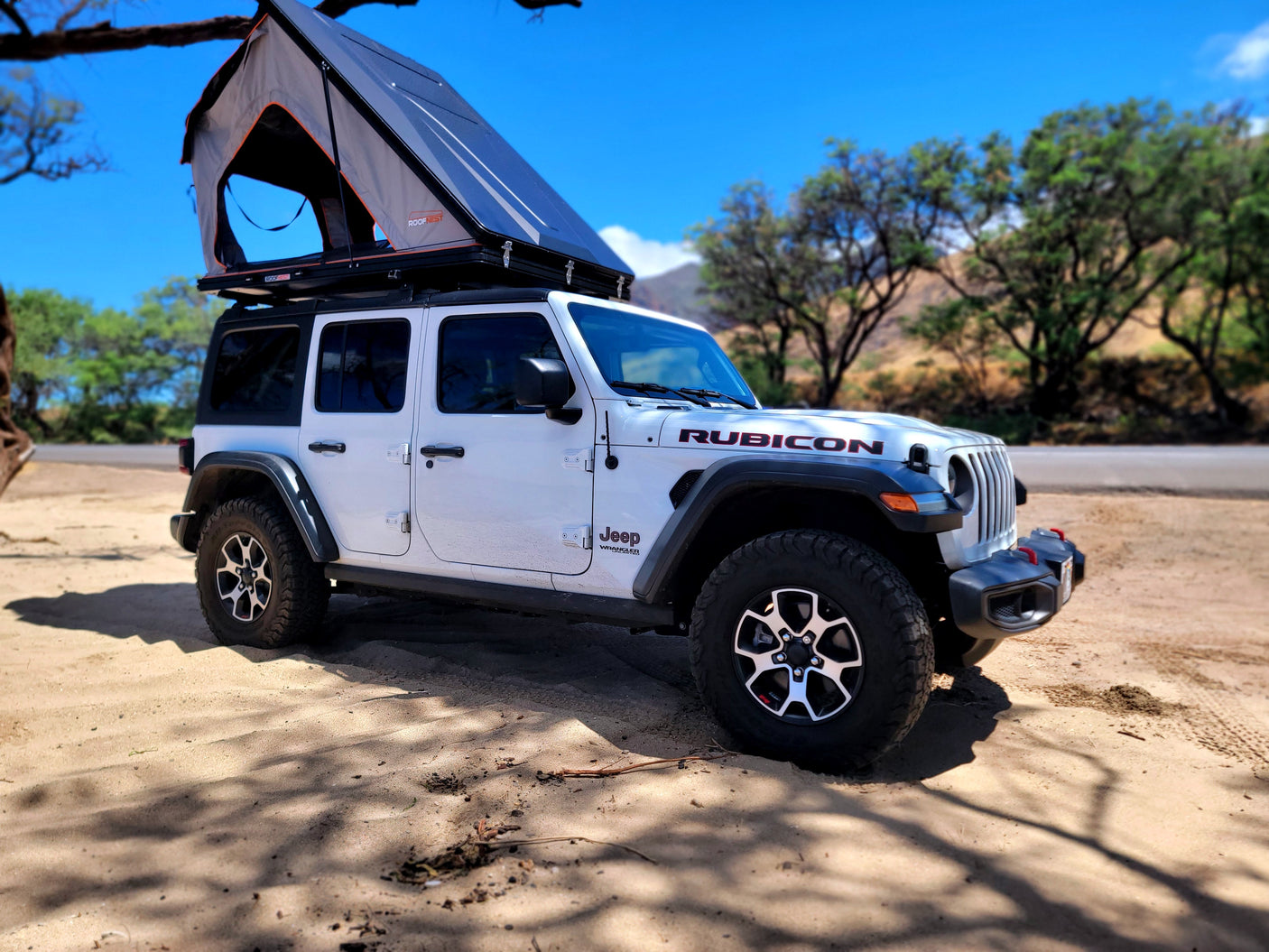 A white Jeep Wrangler Rubicon with an open Roofnest tent, parked on sandy ground. Ideal for your adventurous rv rental.