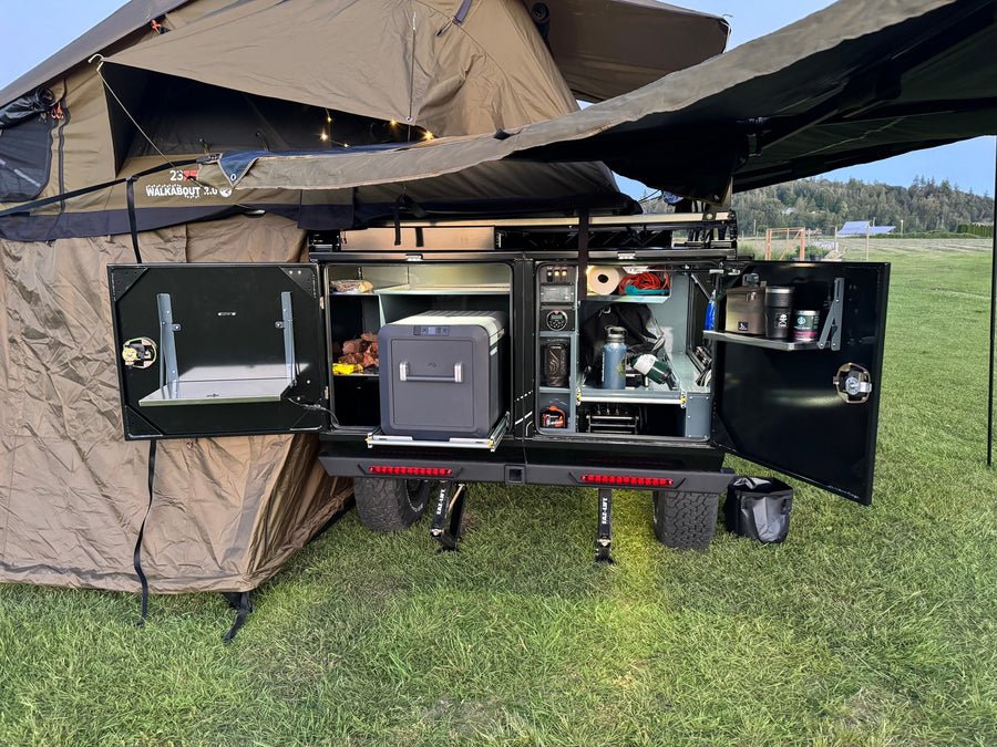 Adventure-ready RV rental setup: an open outdoor kitchen with portable fridge, organized storage, and a deployed rooftop tent.