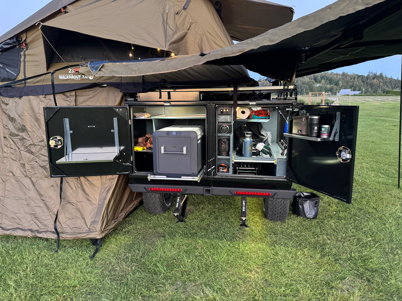 Adventure-ready RV rental setup: an open outdoor kitchen with portable fridge, organized storage, and a deployed rooftop tent.