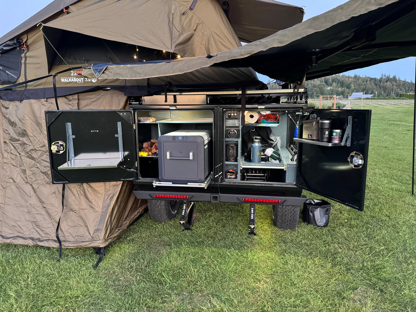 Adventure-ready RV rental setup: an open outdoor kitchen with portable fridge, organized storage, and a deployed rooftop tent.