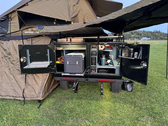Adventure-ready RV rental setup: an open outdoor kitchen with portable fridge, organized storage, and a deployed rooftop tent.