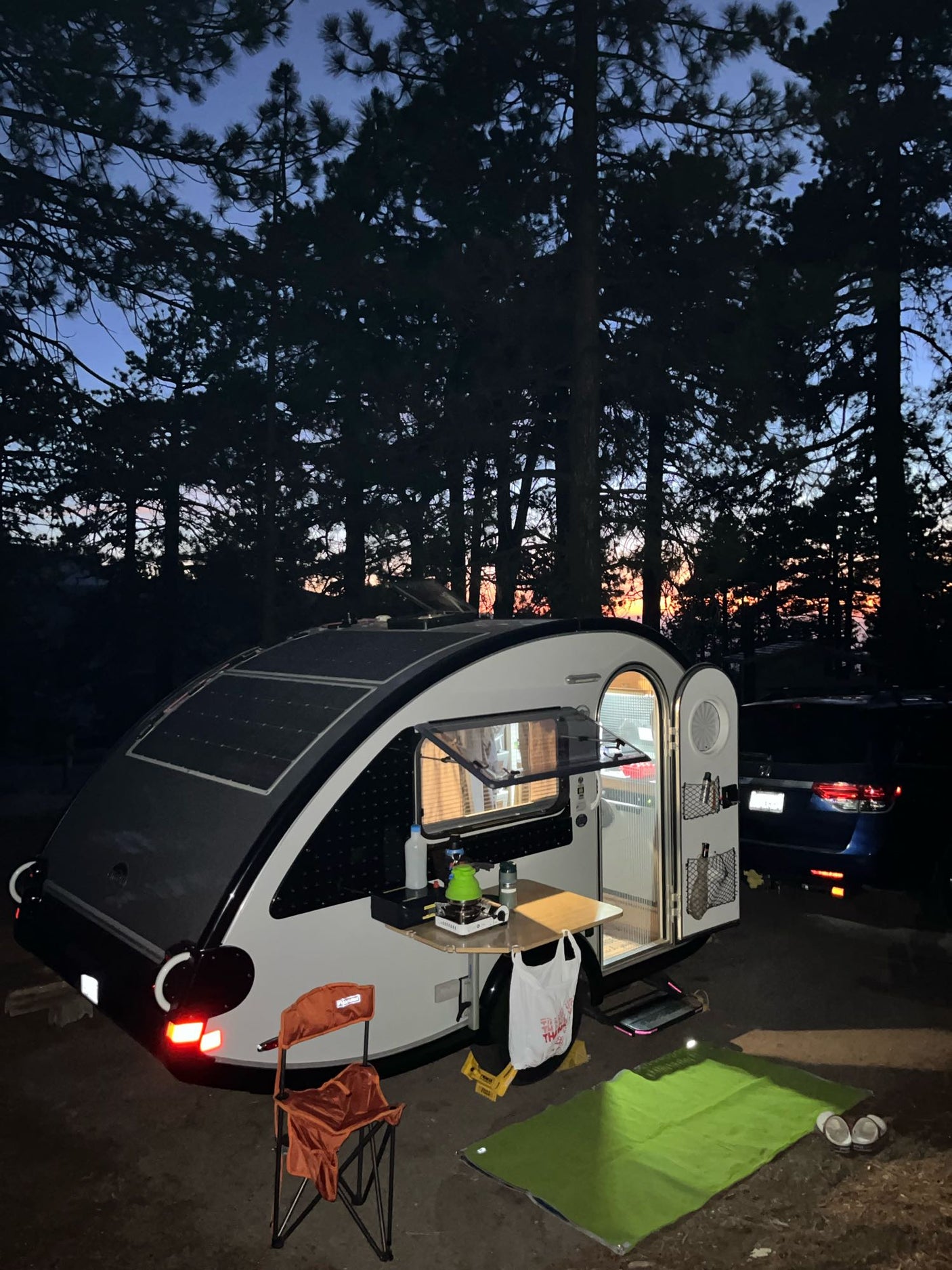 Twilight camping with a modern rv rental teardrop trailer, solar panels & open kitchen in a serene pine forest setting.
