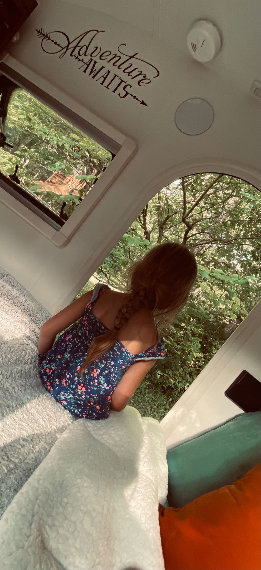 Cozy RV rental view: child looks out at lush trees. "Adventure Awaits" decal on ceiling.
