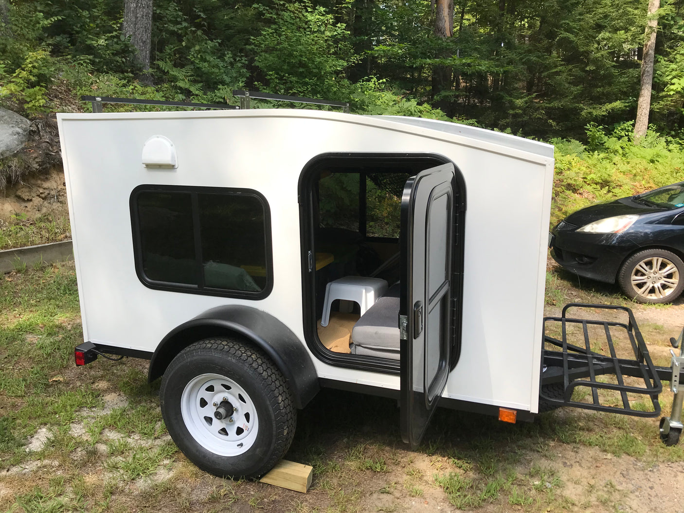 Compact teardrop RV rental in a wooded campsite, featuring an open door and roof rack, perfect for outdoor adventure.