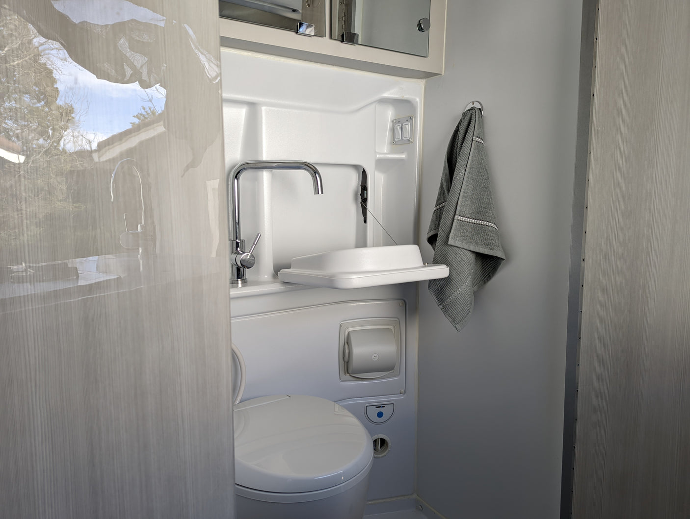 Compact RV rental wet bath, featuring toilet, sink, faucet, and towel. Includes light & fan controls.