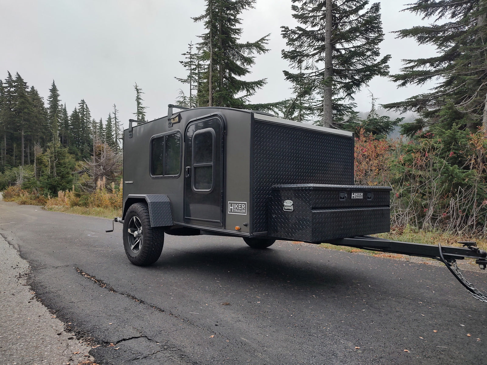 Compact Hiker Trailer RV rental parked on a mountain road, ready for adventure amidst tall evergreen trees.