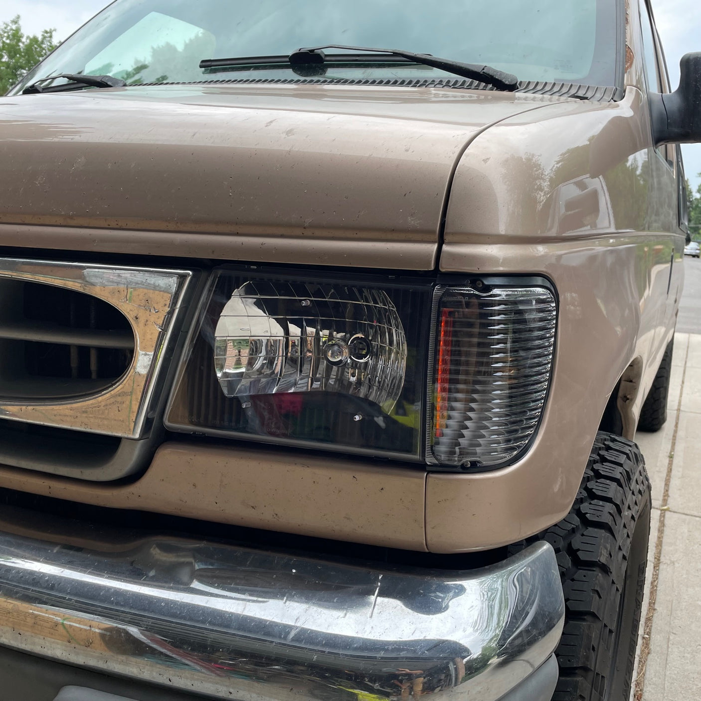 Front close-up of a tan RV rental van, showing the headlight, chrome grille, and rugged tires, ready for adventure.