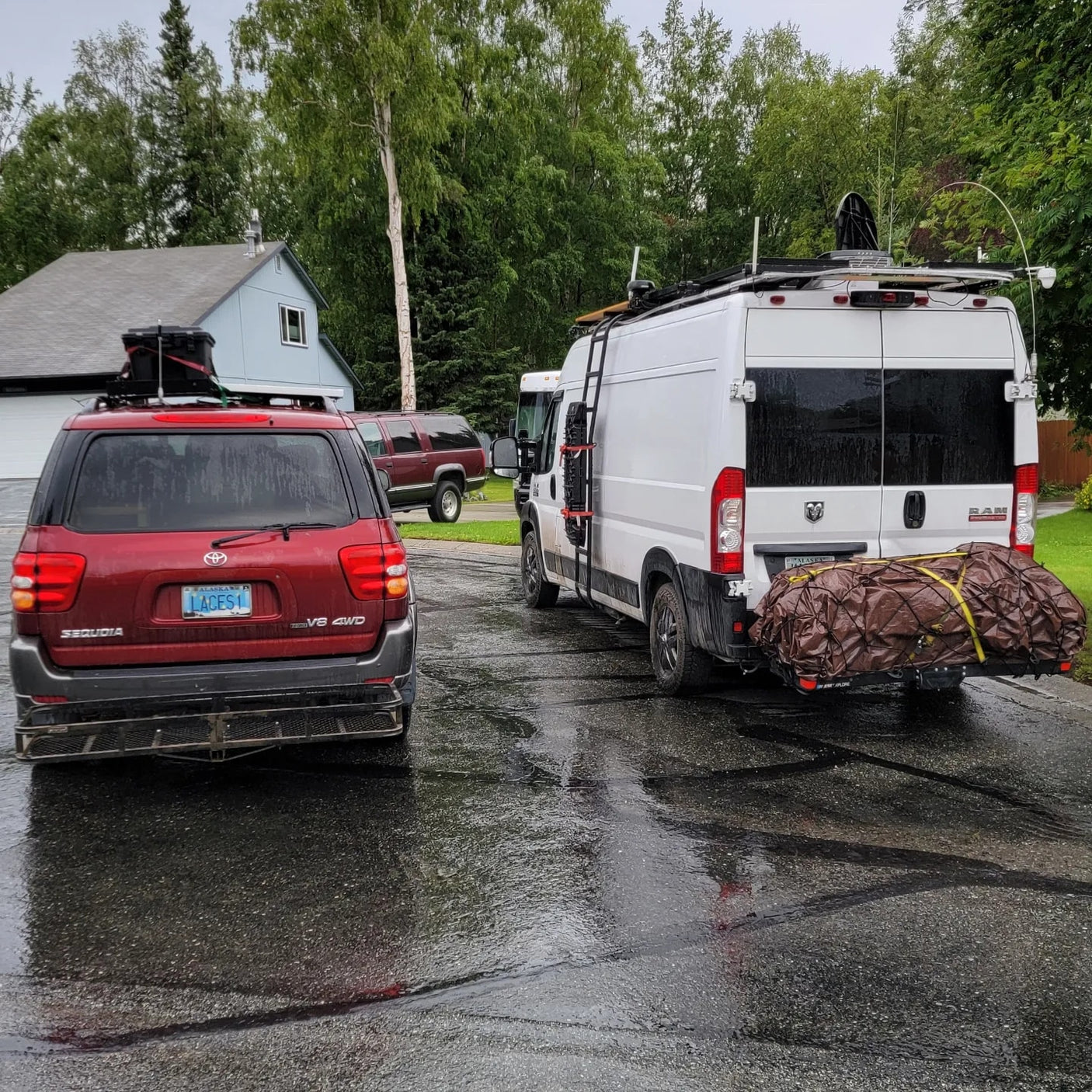 Explore with this **RV rental**! A white customized van & red SUV, loaded with travel gear, parked on a wet street.
