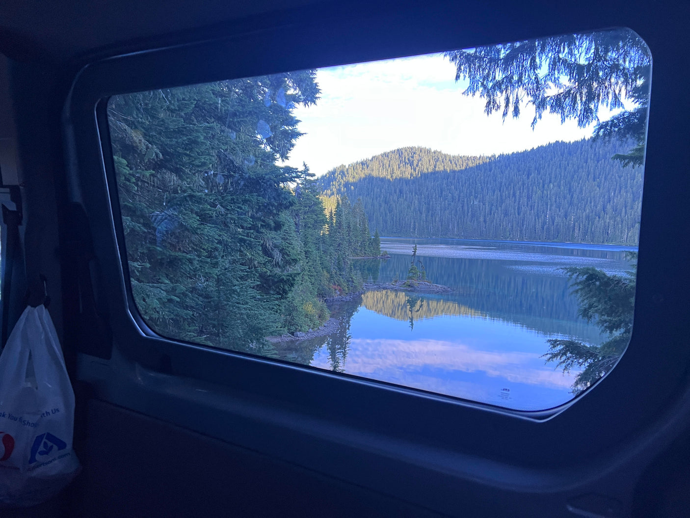 Tranquil lake, evergreen trees, and mountains reflected outside an RV rental window. Perfect nature view.