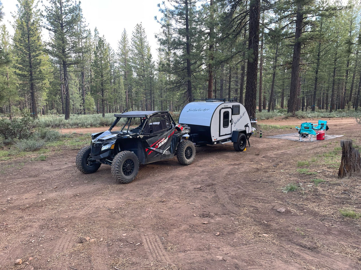 A black off-road UTV is hitched to a white teardrop RV rental camper at a scenic forest campsite