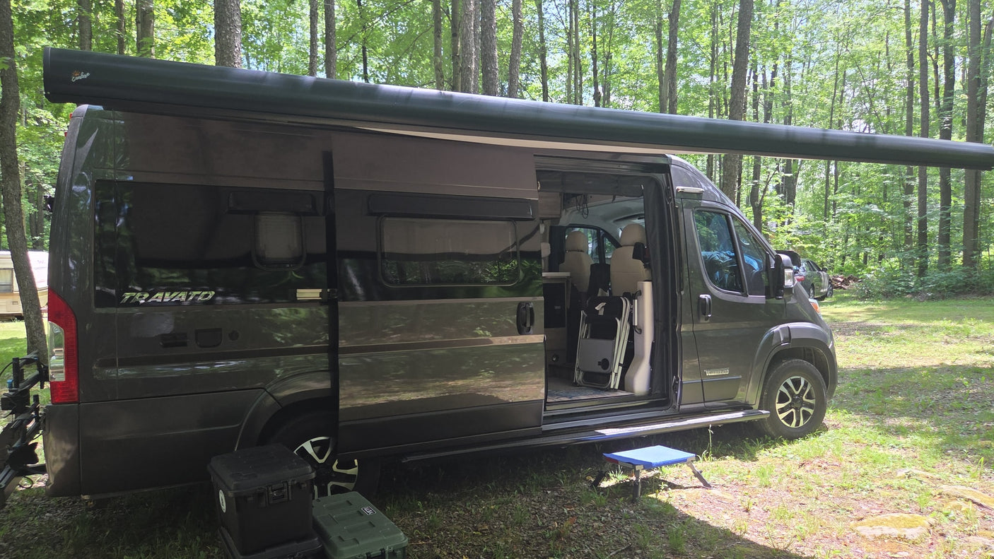 Enjoying the forest in a dark gray Winnebago Travato RV rental with its awning extended and door open, perfect for camping.
