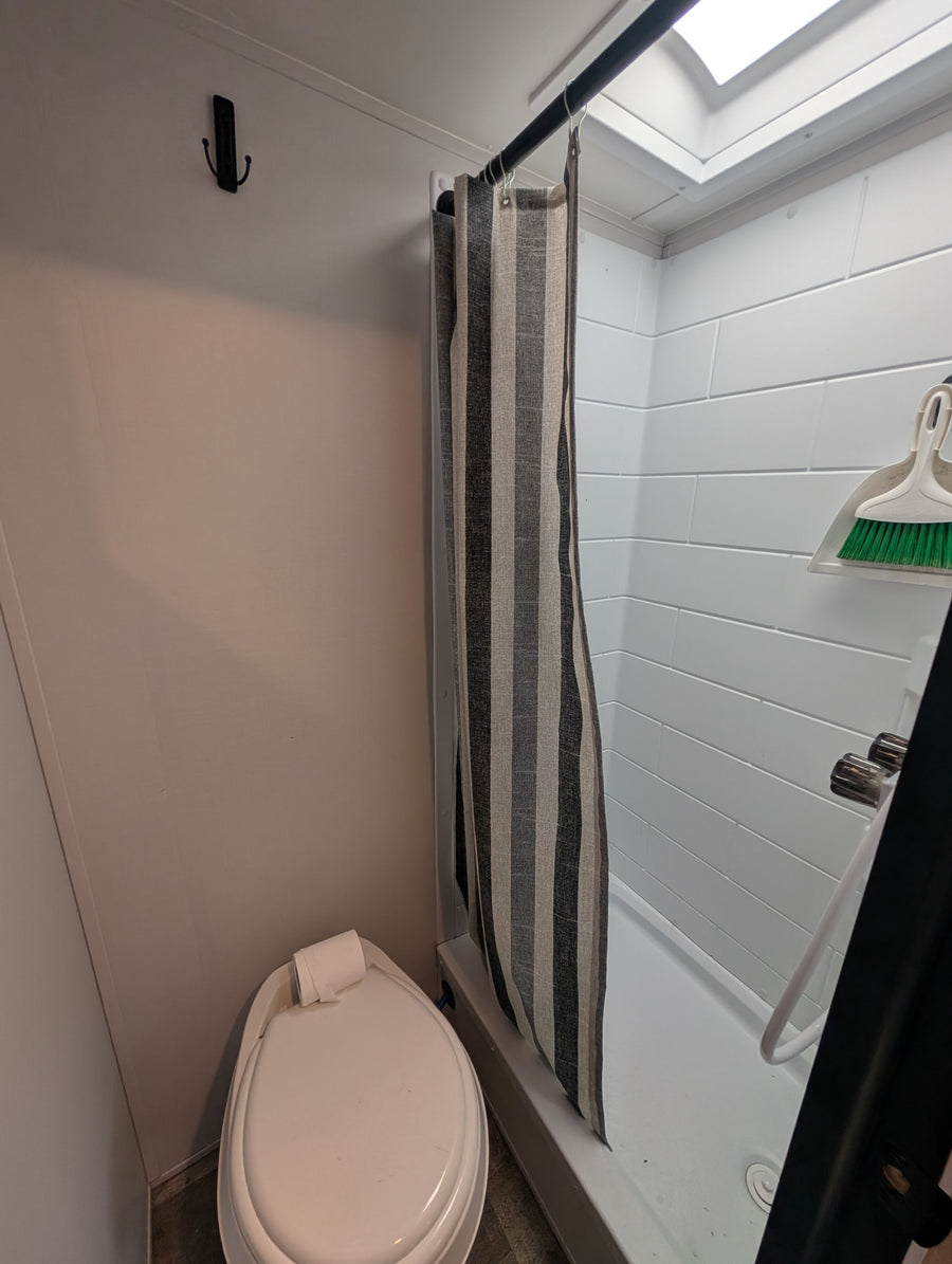 Compact RV rental bathroom: toilet, striped shower, overhead skylight, and cleaning supplies.