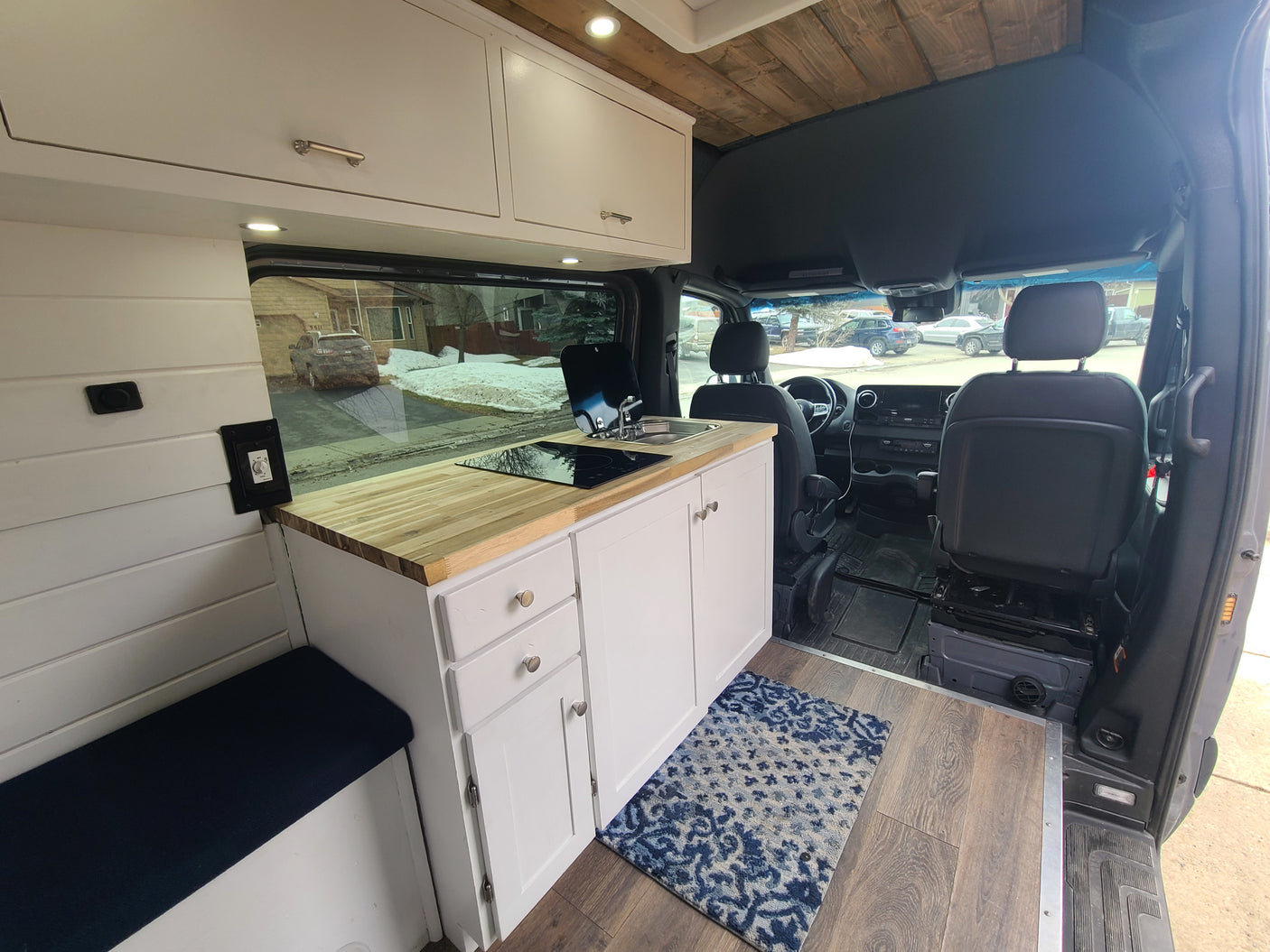 Bright custom RV rental interior: white cabinets, wood counter, cooktop, sink, comfy bench. A modern camper van conversion.