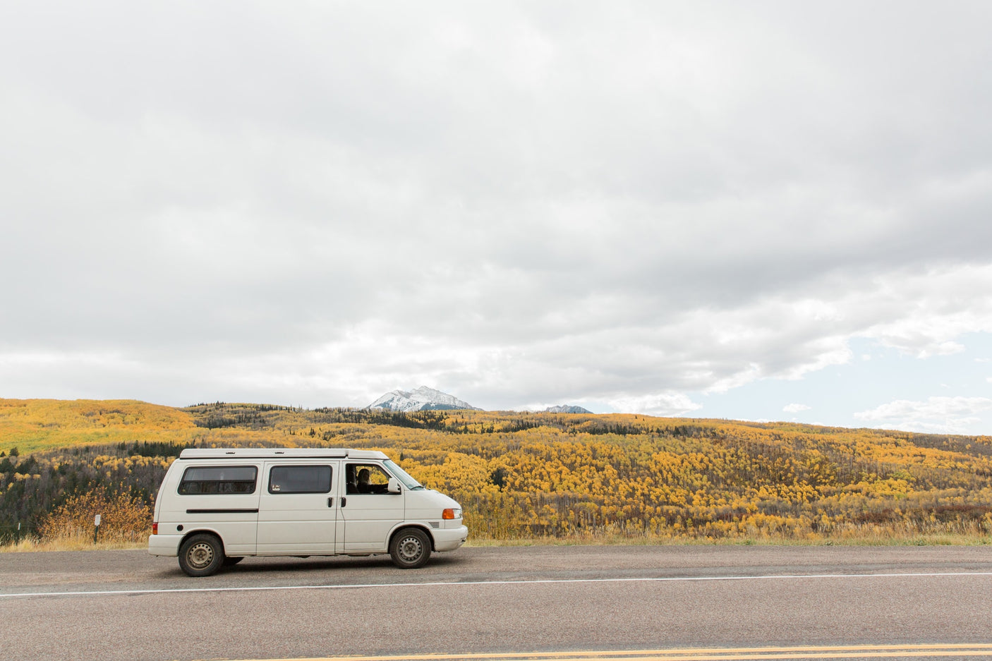 RV rental on a scenic road trip: A white camper van parked alongside a mountain road with vibrant yellow fall foliage and snow-capped peaks under a cloudy sky.