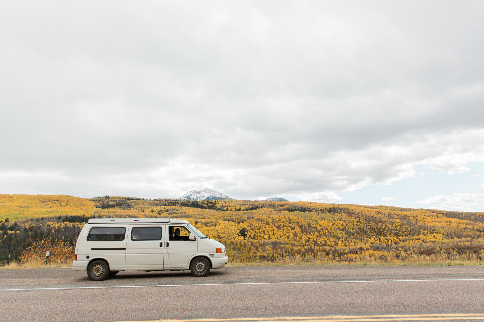 RV rental on a scenic road trip: A white camper van parked alongside a mountain road with vibrant yellow fall foliage and snow-capped peaks under a cloudy sky.