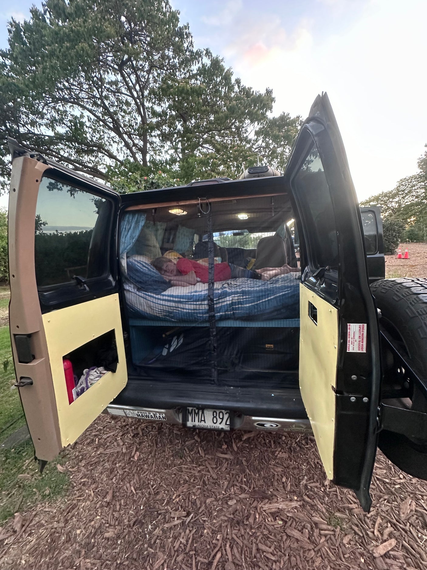 An RV rental camper van with open back doors. A person sleeps on a bed inside, a Hawaii license plate visible.